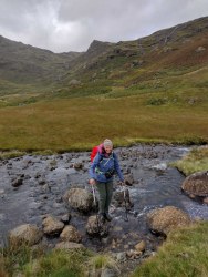 Hiker crossing a rocky beck on stepping stones in a Lake District valley